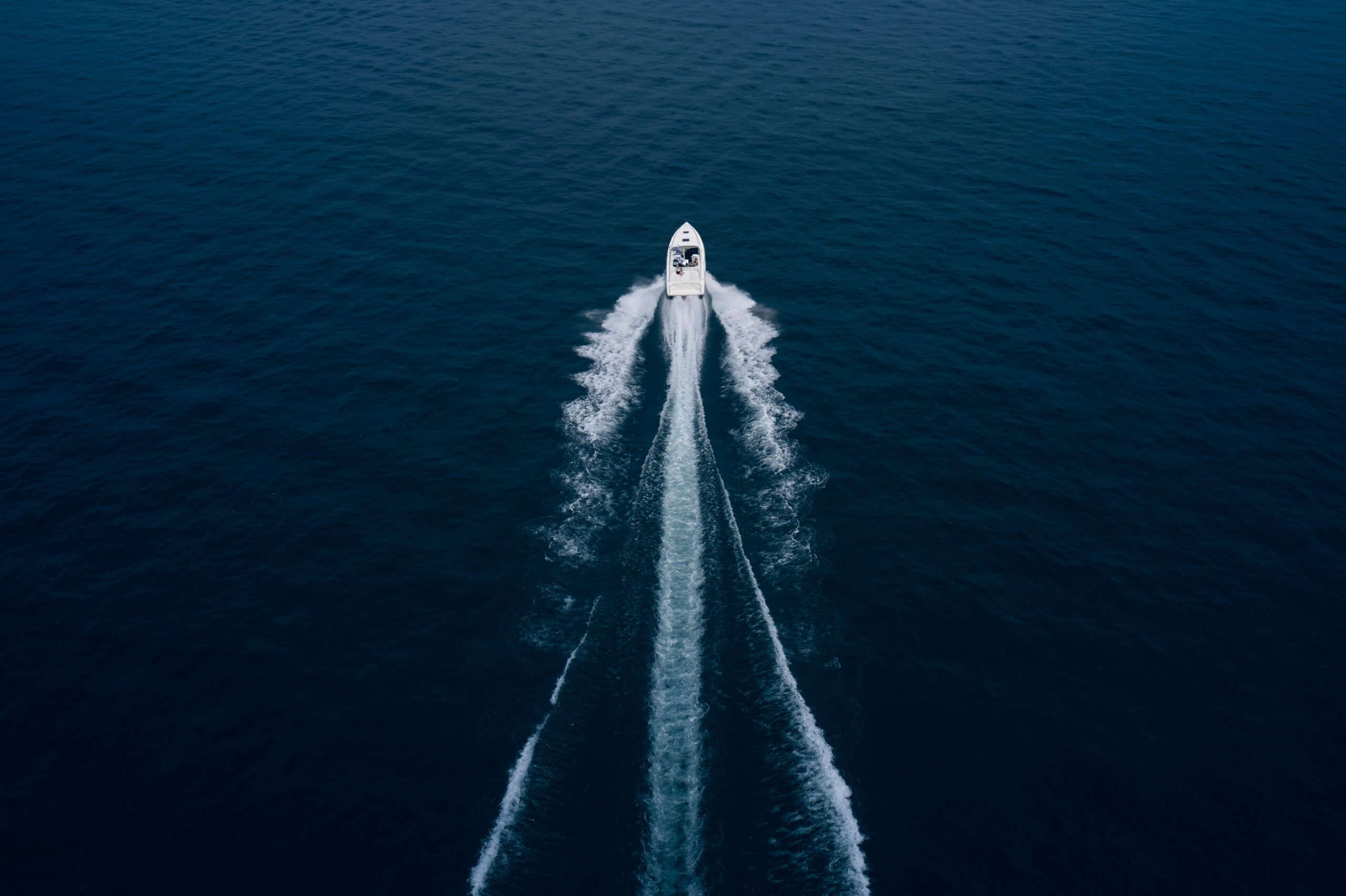 Top-down aerial shot of a white speedboat cutting through deep blue water, leaving symmetrical trails of wake behind.