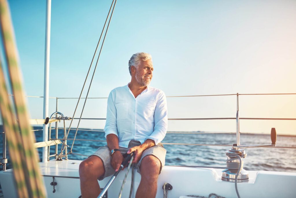 A man with white hair and beard, holding a fishing pole, sitting on the side of a boat on a blue lake.
