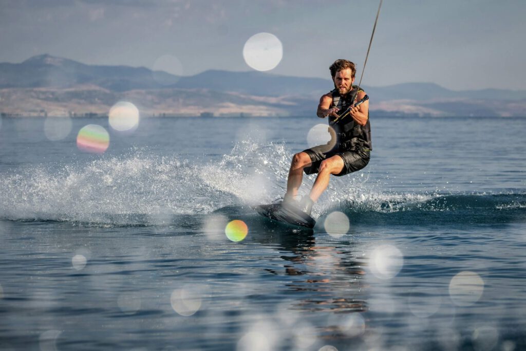 A man in a life vest wakeboarding on a calm lake, kicking up spray with blurred lens flares and mountains in the background. He is making contact with the handle.